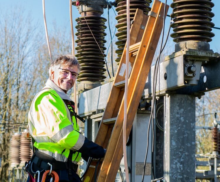 Elektryk w odzieży ostrzegawczej przy drabinie na stacji elektroenergetycznej, w tle izolatory wysokiego napięcia